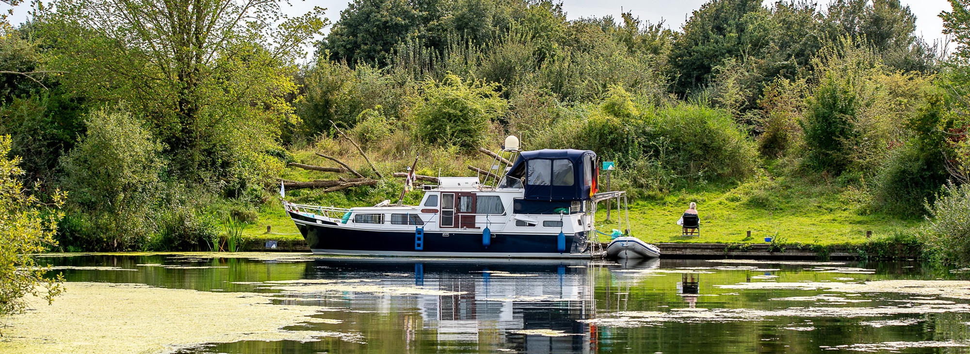 Een motorboot ligt aangemeerd langs een groene oever in een rustige, waterrijke omgeving. Een vrouw zit op een stoel aan de waterkant. Met de boot naar Limburg geniet je van rust, natuur en groene oevers.