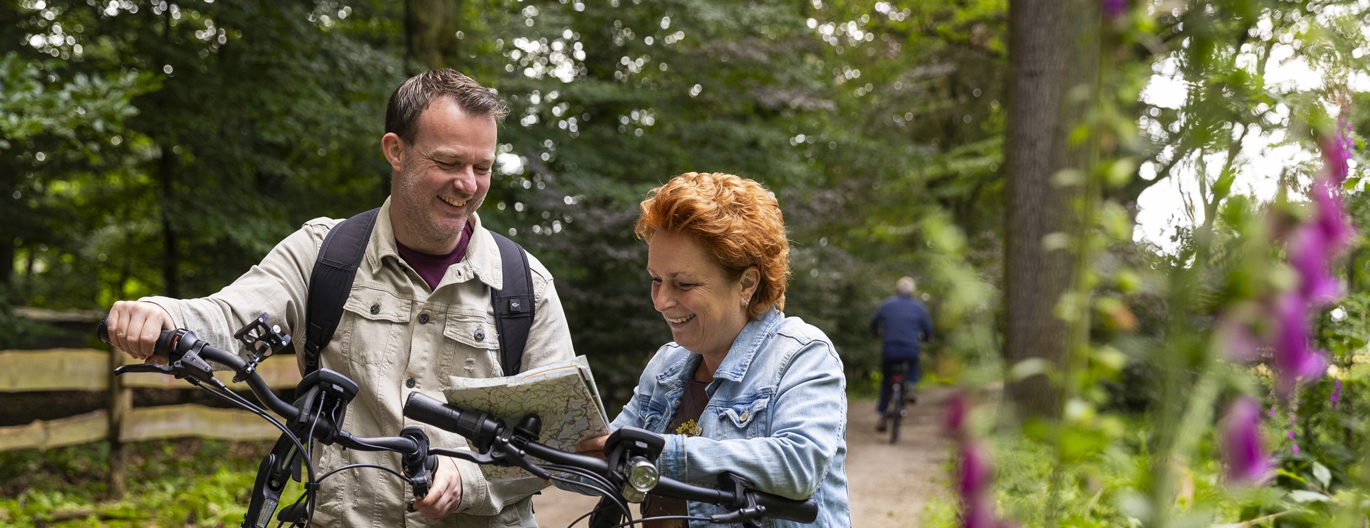 Twee mensen lachen samen terwijl ze met de fiets stilstaan in een bosrijk gebied, kijkend op een fietskaart, met op de achtergrond nog een fietser en kleurrijke bloemen op de voorgrond.