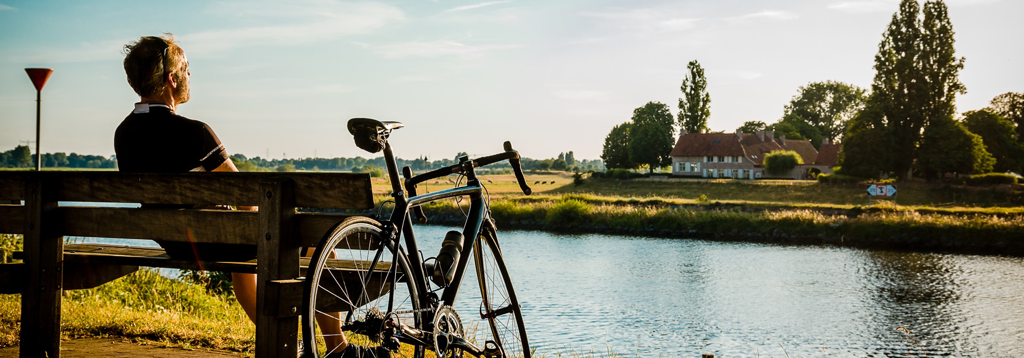 Fietser rust uit op een bankje aan het water in Maasgouw, met uitzicht op de groene oever en een boerderij op de oever tegenover hem.