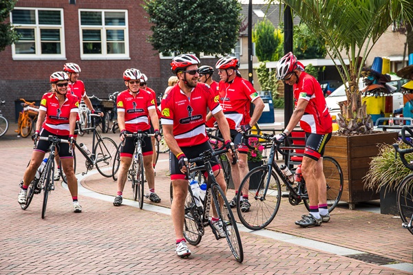 Groep wielrenners in rode sportkleding staat klaar met hun fietsen op een plein in het centrum van Nederweert.