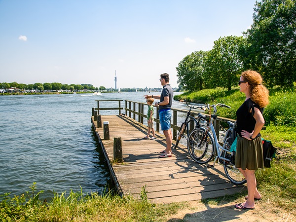 Fietsers staan met hun fietsen aan de oever van de Maas en kijken uit over het water.