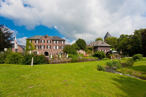 View of the historic centre of Brüggen with its castle, water mill and waterside terrace, surrounded by greenery and clear skies.