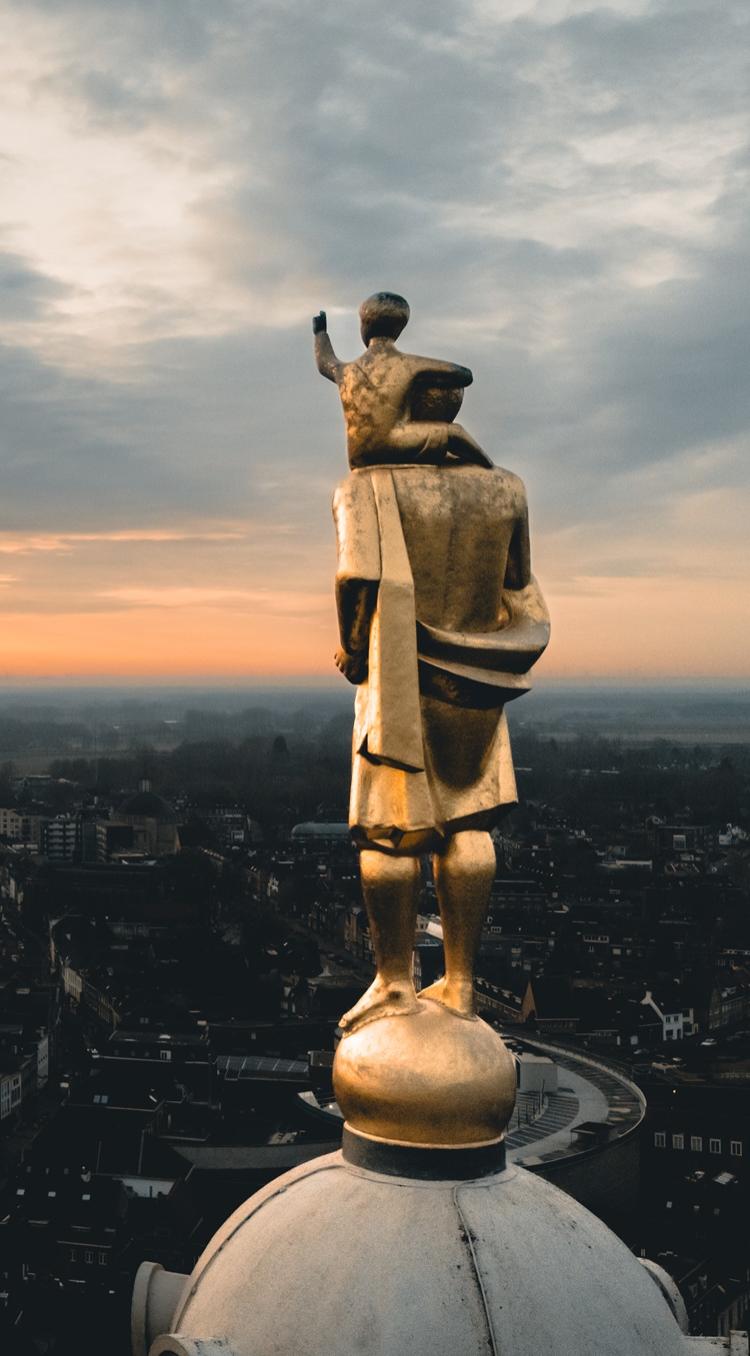 Goudkleurig beeld van Sint-Christoffel met het Christuskind op zijn schouders, uitkijkend over de daken van Roermond bij zonsondergang.