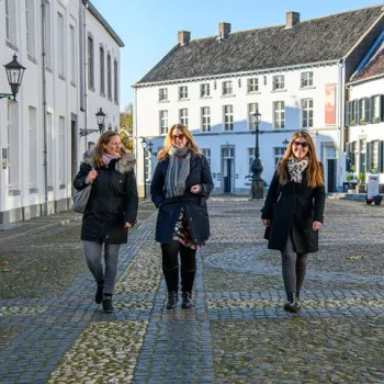 Drie vrouwen in winterkleding wandelen lachend over een zonnig plein met kinderkopjes en witte gebouwen in het centrum van Thorn.