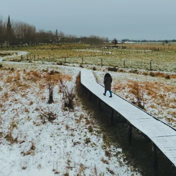 Persoon wandelt over een houten vlonderpad door een besneeuwd winterlandschap bij een dorp met kerktoren in de verte.