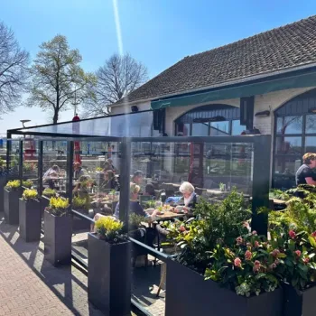 Gasten zitten op het zonnige terras van Gasterie De Knip, omringd door plantenbakken en glazen windschermen.