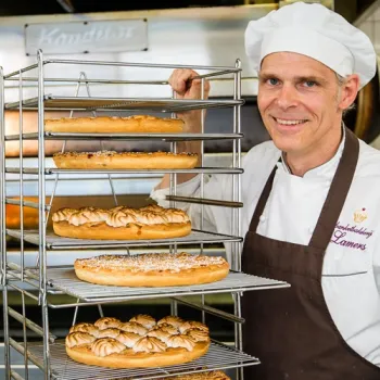 Limburgse vlaaibakker staat naast een rek met versgebakken vlaaien in de bakkerij.